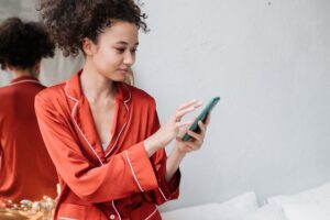 Young woman in red pajamas using smartphone indoors with a mirror reflection, capturing a casual moment.