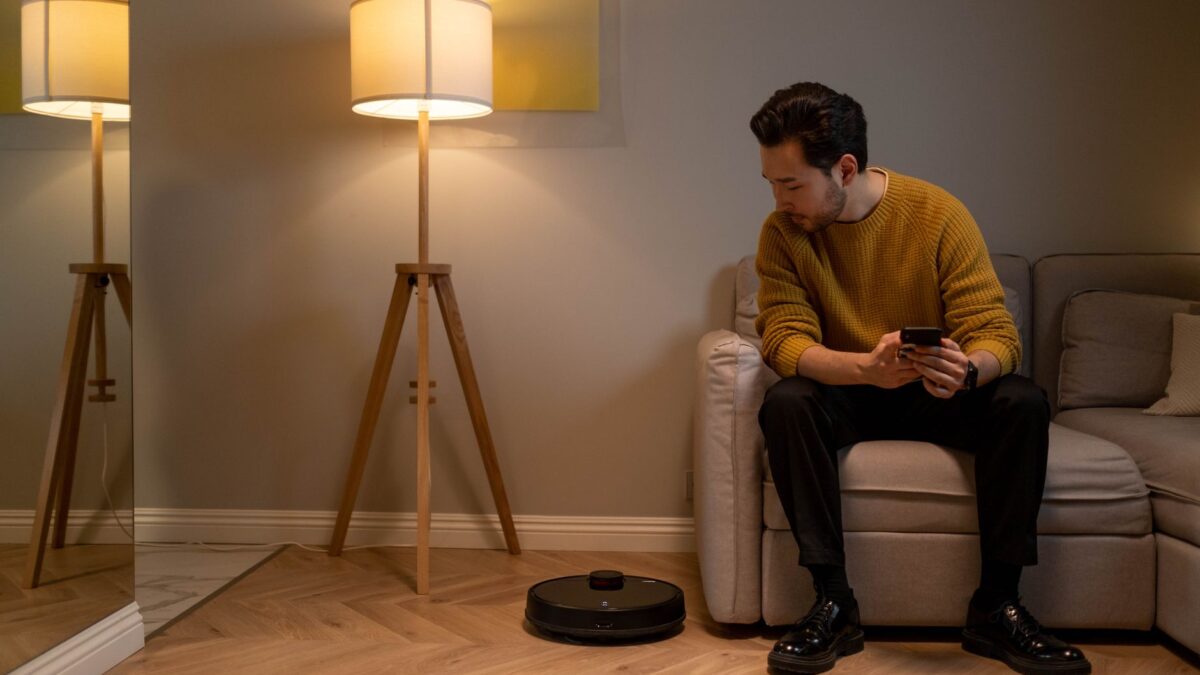 Mann auf Sofa im modernen Wohnzimmer nutzt Smartphone zur Steuerung eines Saugroboters.
