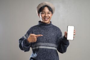 Cheerful young man in glasses and hat pointing at blank smartphone screen, ideal for technology mockups.