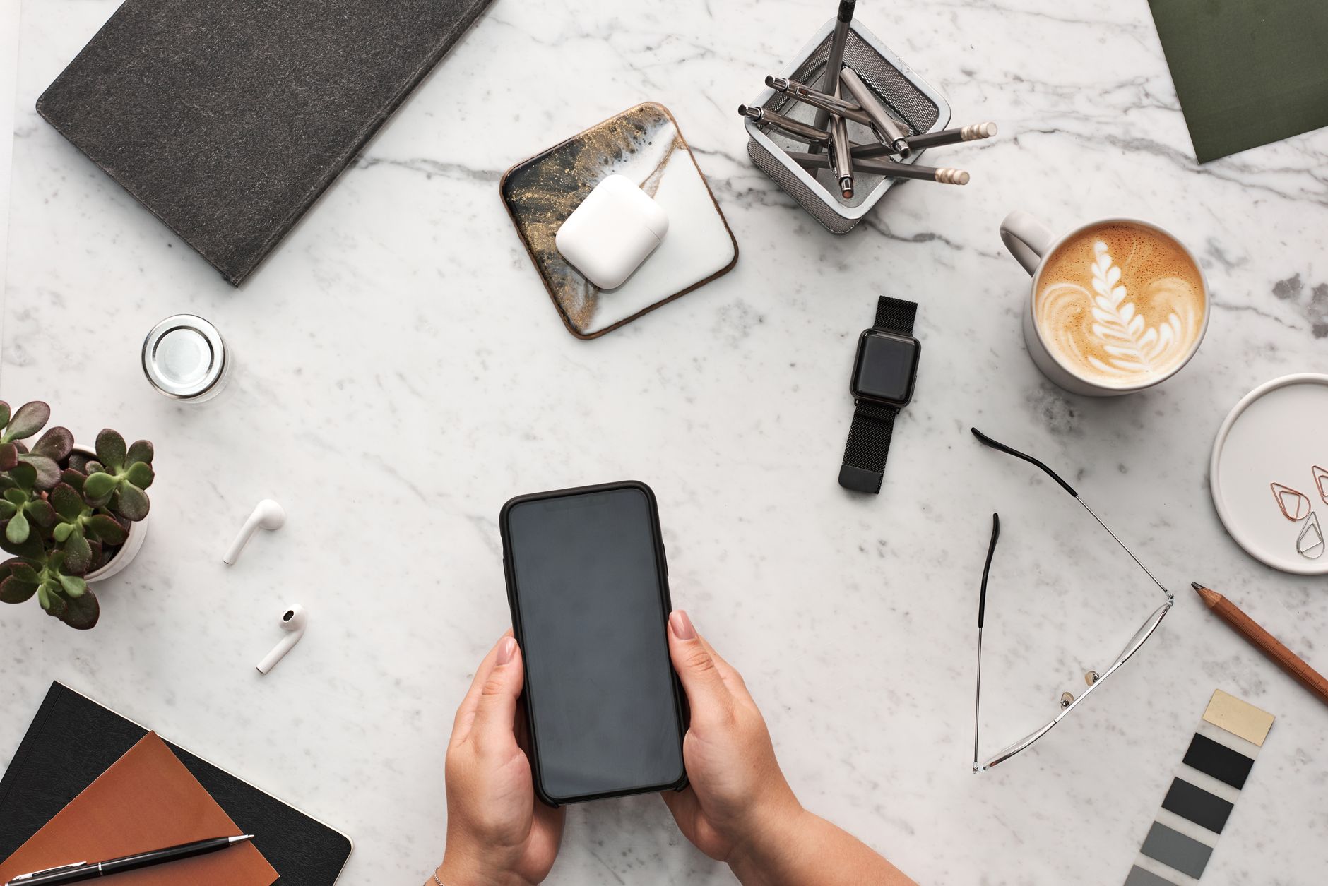 Zwei Hände halten ein Smartphone an einem Schreibtisch mit Kaffeetasse und Zubehör aus der Vogelperspektive.