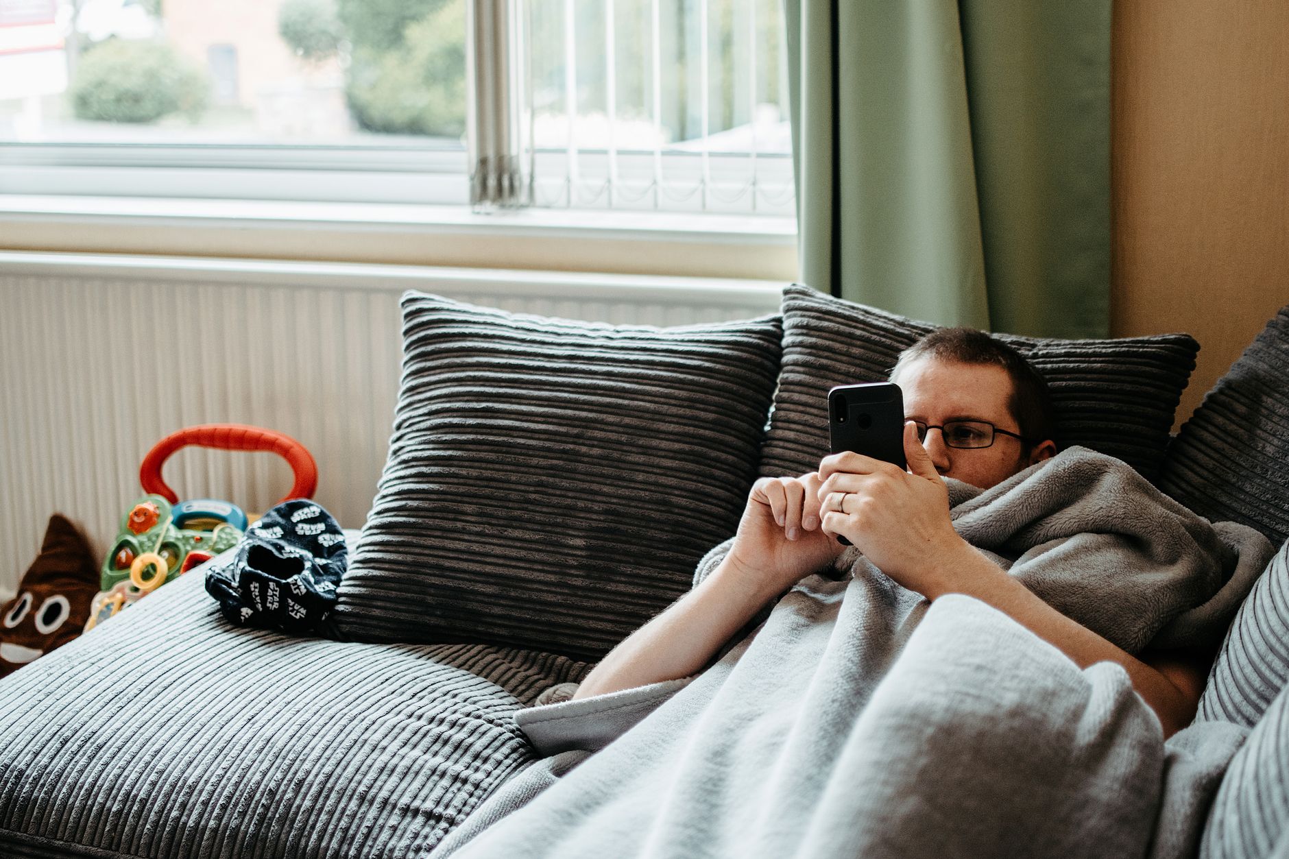 Mann mit Brille liegt unter einer grauen Decke auf einem Sofa und schaut auf ein Smartphone, daneben Kinderspielzeug.