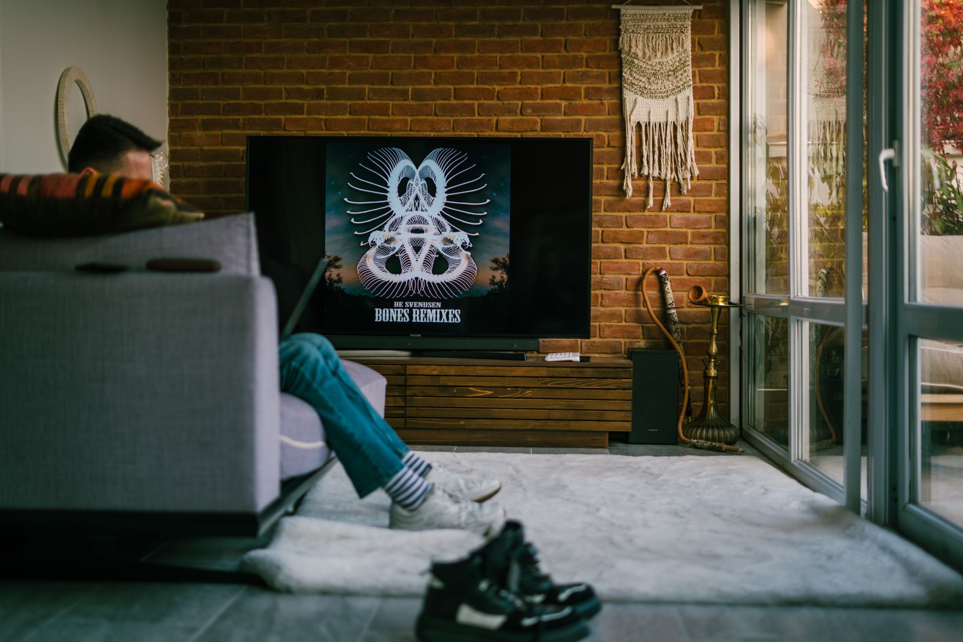 Person sitzt auf einem grauen Sofa und blickt auf einen Fernseher im Wohnzimmer mit Backsteinwand, davor ein heller Teppich.