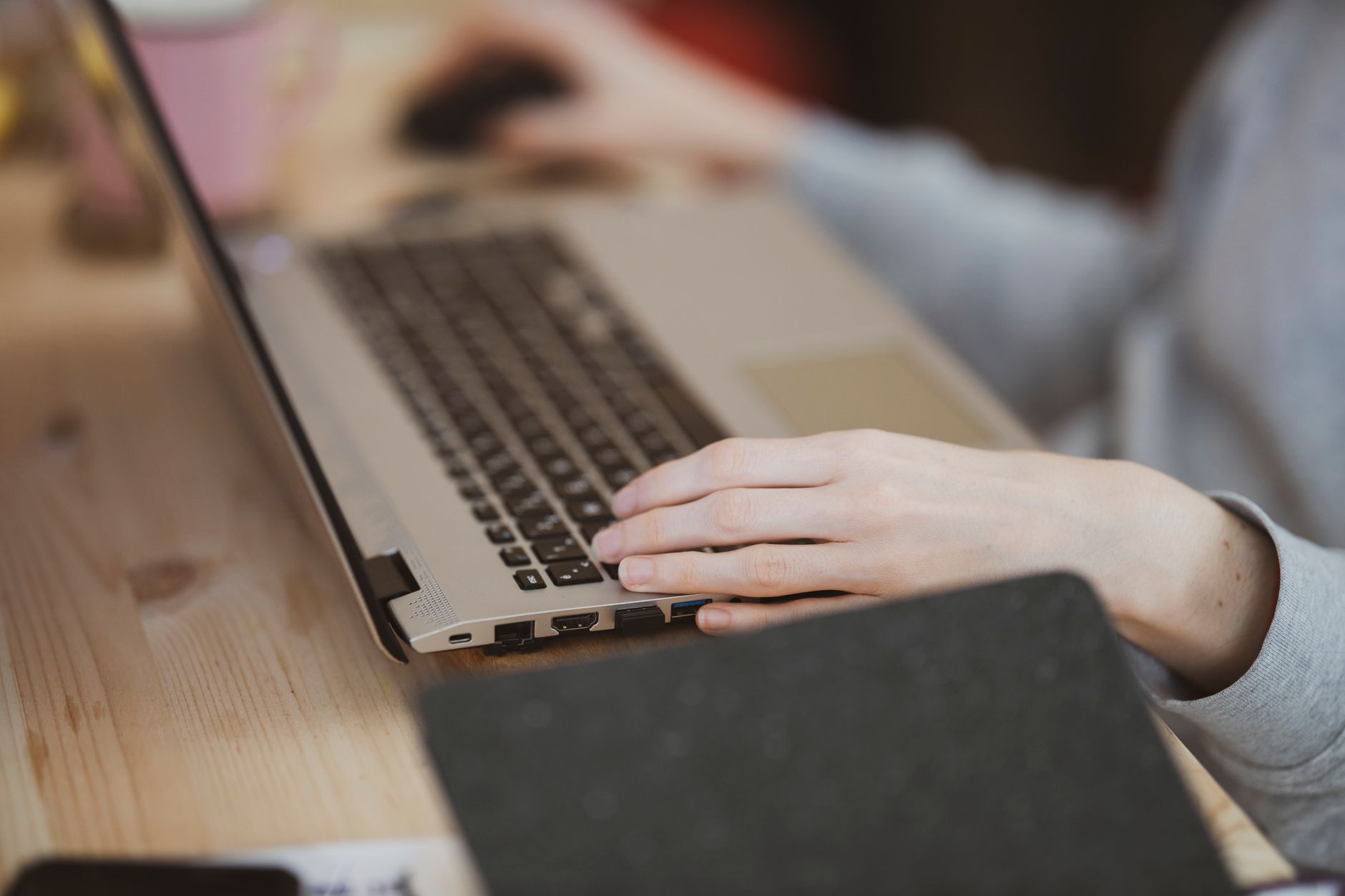 Une main posée sur le clavier d’un ordinateur portable argenté, sur une table en bois, avec une autre main sur une souris en arrière-plan flou.